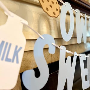 Könnte beinhalten: Ein Banner mit den Wörtern "SWEET" und "MILK" aus weißem und hellblauem Karton ausgeschnitten. Ein Auschnitt eines Kekses ist ebenfalls zu sehen. Das Banner ist auf einem weißen Band aufgereiht, ideal für eine Babyparty oder einen Geburtstag.