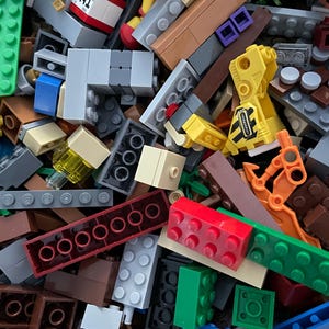 May include: A close-up shot of a pile of colorful plastic building blocks. The blocks are various sizes and shapes, including rectangular bricks, plates, and specialized pieces. Colors include red, green, blue, yellow, gray, and brown. Some blocks have printed details.