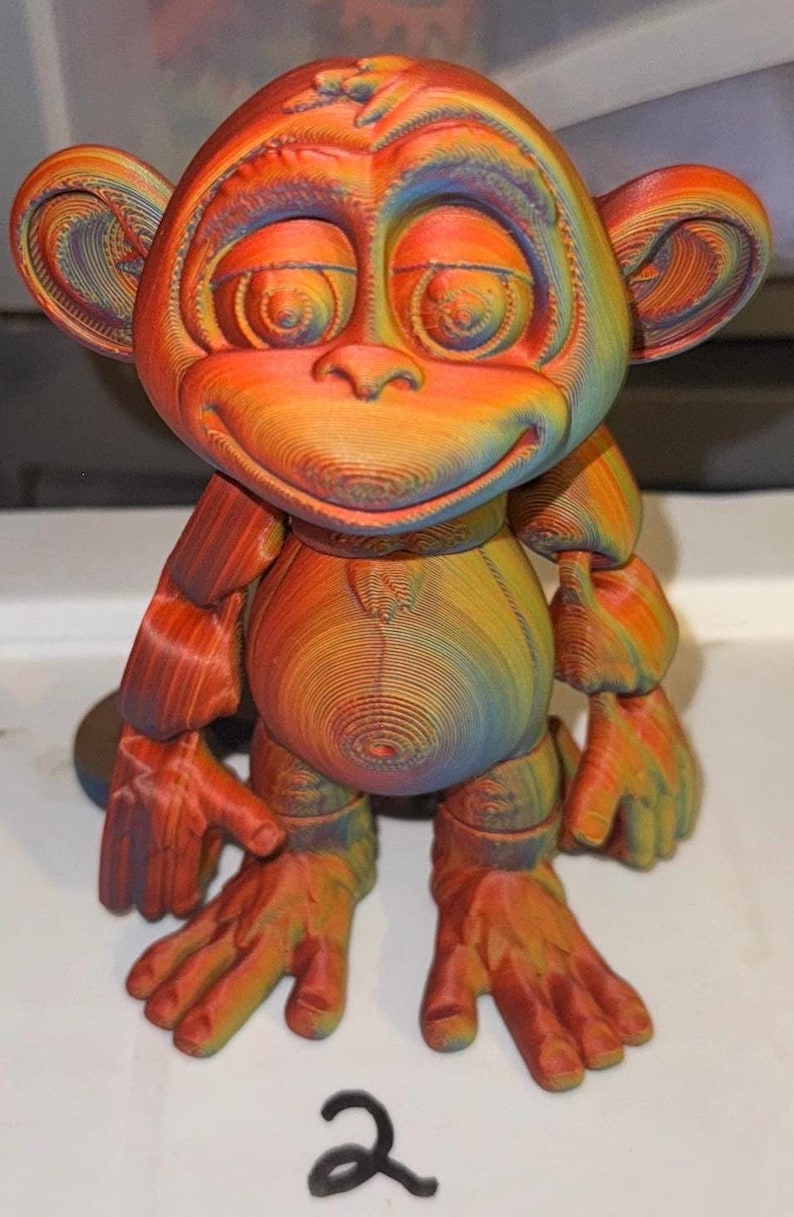 Op de afbeelding: Een 3D-geprinte apenfiguur in regenboogkleuren. De aap heeft een lachend gezicht, grote oren en beweeglijke ledematen. Het cijfer "2" staat in het zwart onderaan. Geschikt voor woondecoratie.