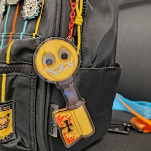 May include: A yellow key charm with a smiling face and red and black details. The charm is attached to a black backpack with a blue and yellow striped pattern.