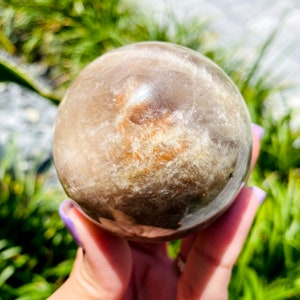 K&ouml;nnte beinhalten: Eine gro&szlig;e, klare, sph&auml;rische Kristallkugel mit einem braunen und wei&szlig;en, wolkigen Inneren. Die Kristallkugel wird in einer Hand gehalten.