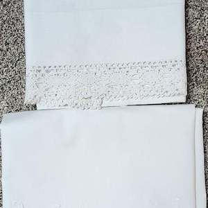 May include: Two folded white pillowcases. The top pillowcase features a wide band of intricate white crochet lace trim. The bottom pillowcase displays a delicate white embroidered floral vine pattern along a scalloped, hemmed edge.