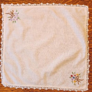 May include: White linen handkerchief with a crocheted border and embroidered floral designs. The handkerchief features two bouquets of flowers, one in the top left corner and one in the bottom right corner.