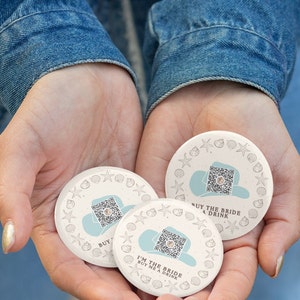 May include: Three round white paper coasters with a blue and white design. Each coaster features a QR code, a seashell border, and the text "Buy the Bride a Drink" or "I'm the Bride Buy Me a Drink".