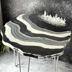 May include: A black and white agate epoxy resin table with a clear crystal cluster on top. The table has a unique, organic shape and is supported by four hairpin legs.