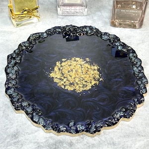May include: A round, dark blue resin tray with a gold leaf design. The tray has a scalloped edge and is perfect for holding small items like jewelry or perfume bottles.
