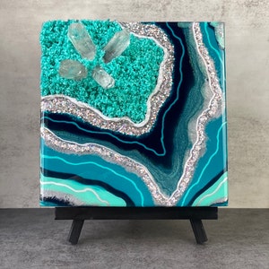May include: A small, square canvas painting with a blue and green agate design. The painting features silver glitter and clear quartz crystals embedded in the resin. The canvas is mounted on a small black easel.