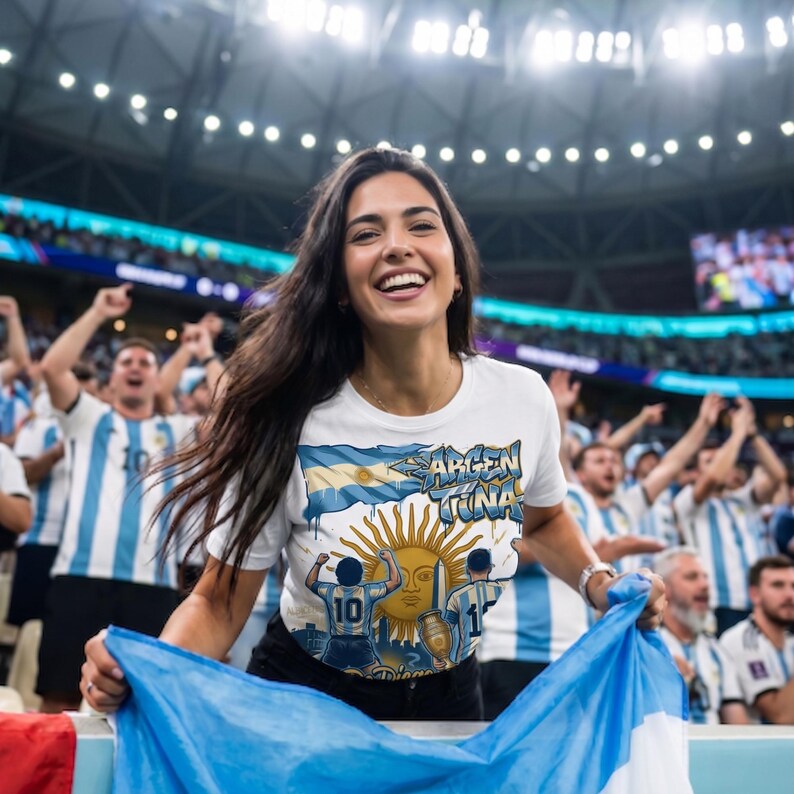 May include: A white t-shirt featuring a graphic print of the Argentine flag, the word Argentina, a golden sun, and two figures wearing number 10 jerseys, one holding a trophy, set against a backdrop of a crowded stadium with fans in blue and white.
