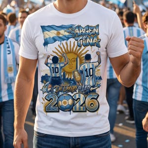 May include: A white t-shirt featuring a graphic print of the Argentine flag, a sun emblem, and two football players wearing number 10 jerseys. The design includes the text "ARGENTINA", "De Diego a Leo", and "2026" alongside a football illustration.