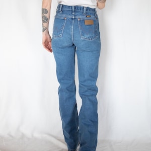 Puede incluir: Vaqueros de mezclilla azul de talle alto con corte recto, dise&ntilde;o cl&aacute;sico de cinco bolsillos y un parche de cuero marr&oacute;n en el bolsillo trasero derecho con el nombre de la marca Wrangler visible en texto negro. Los vaqueros tienen remaches cobrizos.