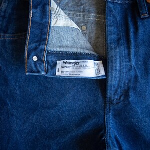 May include: Dark blue denim jeans featuring a high-waisted design with a button fly closure. A white interior label displays the Wrangler brand name, the style number 936DEN, and the size 30 x 36, along with care instructions and fabric details.
