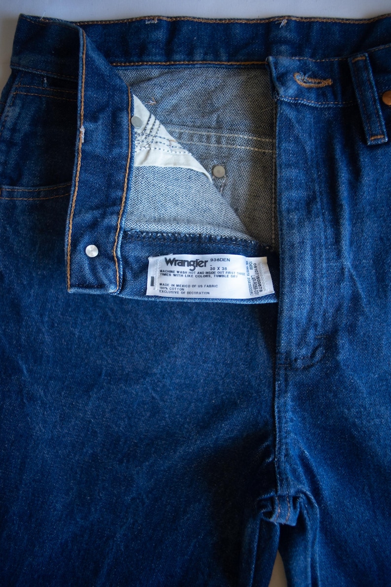 May include: Dark blue denim jeans featuring a high-waisted design with a button fly closure. A white interior label displays the Wrangler brand name, the style number 936DEN, and the size 30 x 36, along with care instructions and fabric details.