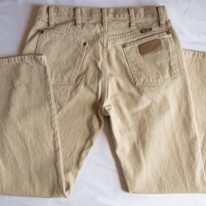 Op de afbeelding: Een beige spijkerbroek van denim, gezien vanaf de achterkant, met een hoge taille en riemlussen, twee achterzakken met koperkleurige klinknagels en een bruine leren patch op de rechterzak met de merknaam Wrangler duidelijk zichtbaar.