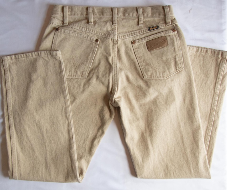 Op de afbeelding: Een beige spijkerbroek van denim, gezien vanaf de achterkant, met een hoge taille en riemlussen, twee achterzakken met koperkleurige klinknagels en een bruine leren patch op de rechterzak met de merknaam Wrangler duidelijk zichtbaar.