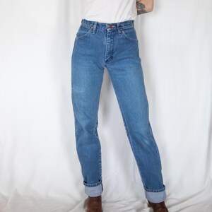 May include: High-waisted blue denim jeans featuring a classic five-pocket design, a metal button closure, and a zip fly. The jeans have a straight-leg cut with visible stitching along the seams and are cuffed at the bottom hem, paired with a white top.