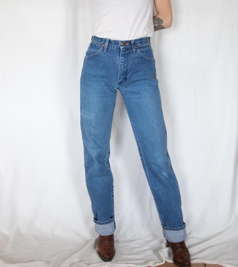 May include: High-waisted blue denim jeans featuring a classic five-pocket design, a metal button closure, and a zip fly. The jeans have a straight-leg cut with visible stitching along the seams and are cuffed at the bottom hem, paired with a white top.