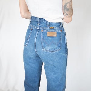 Puede incluir: Vaqueros de mezclilla azul de talle alto con dise&ntilde;o cl&aacute;sico de cinco bolsillos, remaches en tono cobre y un parche de cuero marr&oacute;n en el bolsillo trasero con el nombre de la marca Wrangler. El pantal&oacute;n tiene costuras visibles.