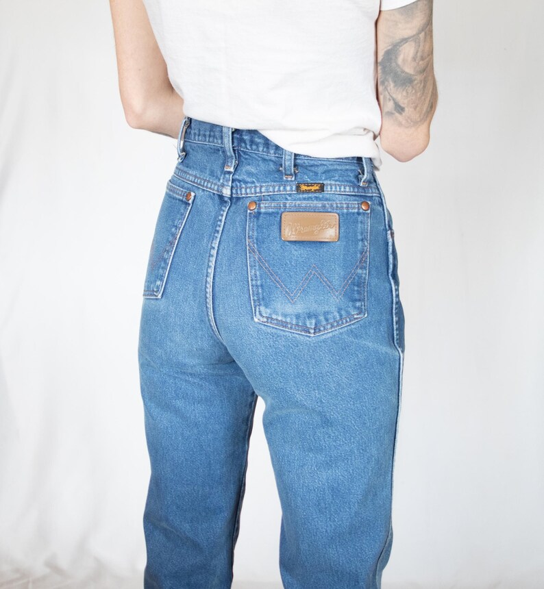 Puede incluir: Vaqueros de mezclilla azul de talle alto con dise&ntilde;o cl&aacute;sico de cinco bolsillos, remaches en tono cobre y un parche de cuero marr&oacute;n en el bolsillo trasero con el nombre de la marca Wrangler. El pantal&oacute;n tiene costuras visibles.