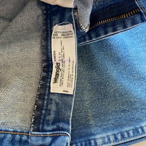 Puede incluir: Vaqueros de mezclilla azul con una etiqueta de cuidado blanca en el interior que muestra el logotipo de Wrangler, la talla 31 x 36 e instrucciones de lavado. La etiqueta indica 100% algod&oacute;n, hecho en M&eacute;xico con tela de EE. UU., RN 130273 y 936DEN.