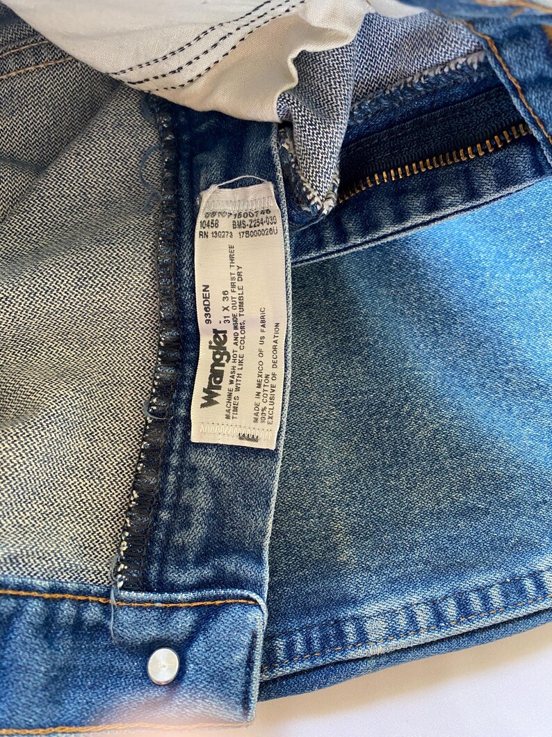 Puede incluir: Vaqueros de mezclilla azul con una etiqueta de cuidado blanca en el interior que muestra el logotipo de Wrangler, la talla 31 x 36 e instrucciones de lavado. La etiqueta indica 100% algod&oacute;n, hecho en M&eacute;xico con tela de EE. UU., RN 130273 y 936DEN.