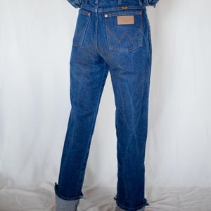 May include: A pair of dark blue denim jeans featuring a high-waisted fit, a leather-like patch on the back pocket, and the Wrangler brand logo. The jeans have a classic five-pocket design with copper-coloured rivets and a cuffed hem at the ankles.