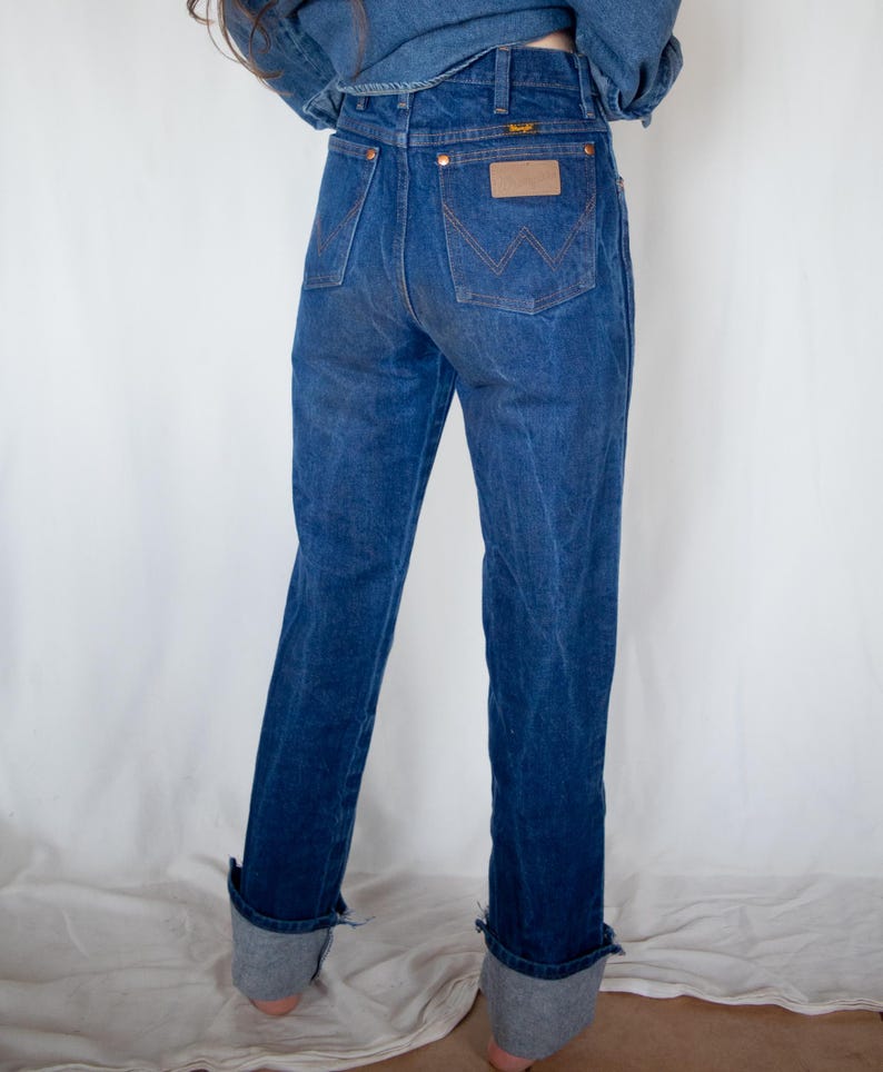 May include: A pair of dark blue denim jeans featuring a high-waisted fit, a leather-like patch on the back pocket, and the Wrangler brand logo. The jeans have a classic five-pocket design with copper-coloured rivets and a cuffed hem at the ankles.