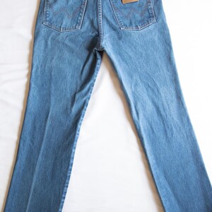 Puede incluir: Un par de vaqueros de mezclilla en tono azul medio con un dise&ntilde;o cl&aacute;sico de cinco bolsillos, cintura alta con trabillas para cintur&oacute;n y un parche de cuero color canela en el bolsillo trasero derecho con la marca Wrangler en relieve.