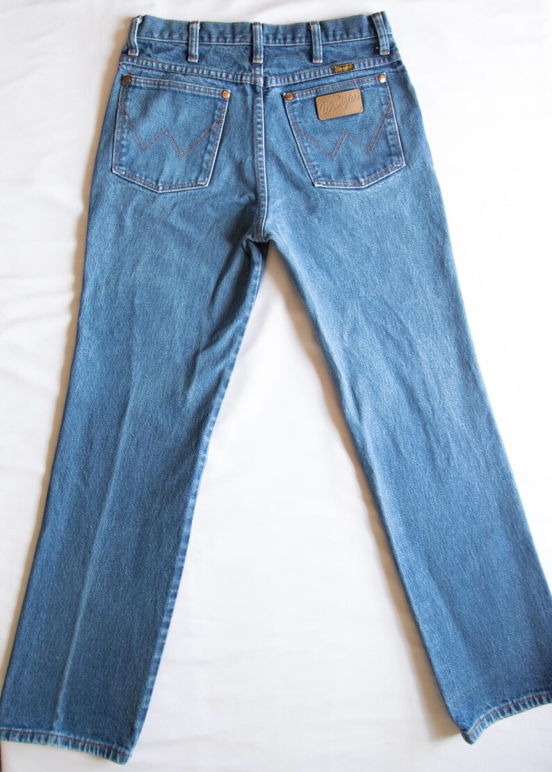 Puede incluir: Un par de vaqueros de mezclilla en tono azul medio con un dise&ntilde;o cl&aacute;sico de cinco bolsillos, cintura alta con trabillas para cintur&oacute;n y un parche de cuero color canela en el bolsillo trasero derecho con la marca Wrangler en relieve.