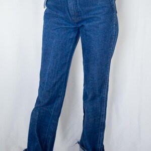 May include: High-waisted dark blue denim jeans featuring a straight-leg cut, a button fly closure with a metallic button, copper-toned rivets at the pockets, and wide, folded cuffs at the bottom hem, paired with a matching blue denim jacket.