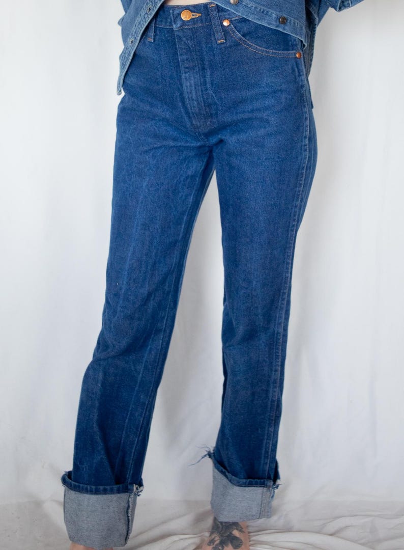 May include: High-waisted dark blue denim jeans featuring a straight-leg cut, a button fly closure with a metallic button, copper-toned rivets at the pockets, and wide, folded cuffs at the bottom hem, paired with a matching blue denim jacket.
