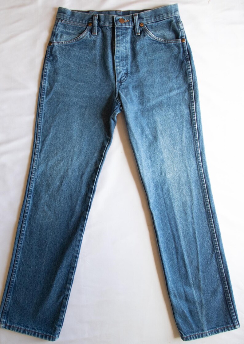 Puede incluir: Un par de pantalones vaqueros de mezclilla en tono azul medio con un dise&ntilde;o cl&aacute;sico de cinco bolsillos, cierre de botones y remaches de color cobre en las esquinas de los bolsillos. Corte recto con costuras visibles en los laterales.