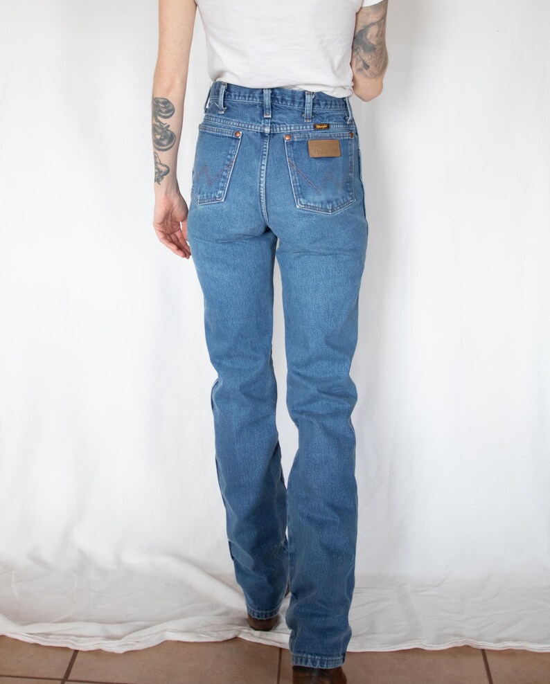 Puede incluir: Vaqueros de mezclilla azul de talle alto con corte recto, dise&ntilde;o cl&aacute;sico de cinco bolsillos y un parche de cuero marr&oacute;n en el bolsillo trasero derecho con el nombre de la marca Wrangler visible en texto negro. Los vaqueros tienen remaches cobrizos.
