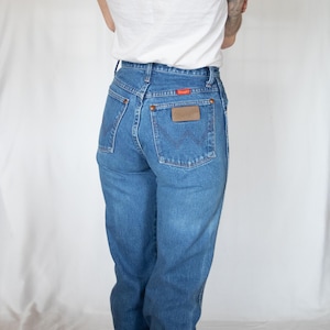 May include: High-waisted blue denim jeans featuring a classic five-pocket design, copper-toned rivets, and a brown leather patch on the back pocket. The back pocket displays a signature W-shaped stitching pattern and a small red Wrangler brand tag.