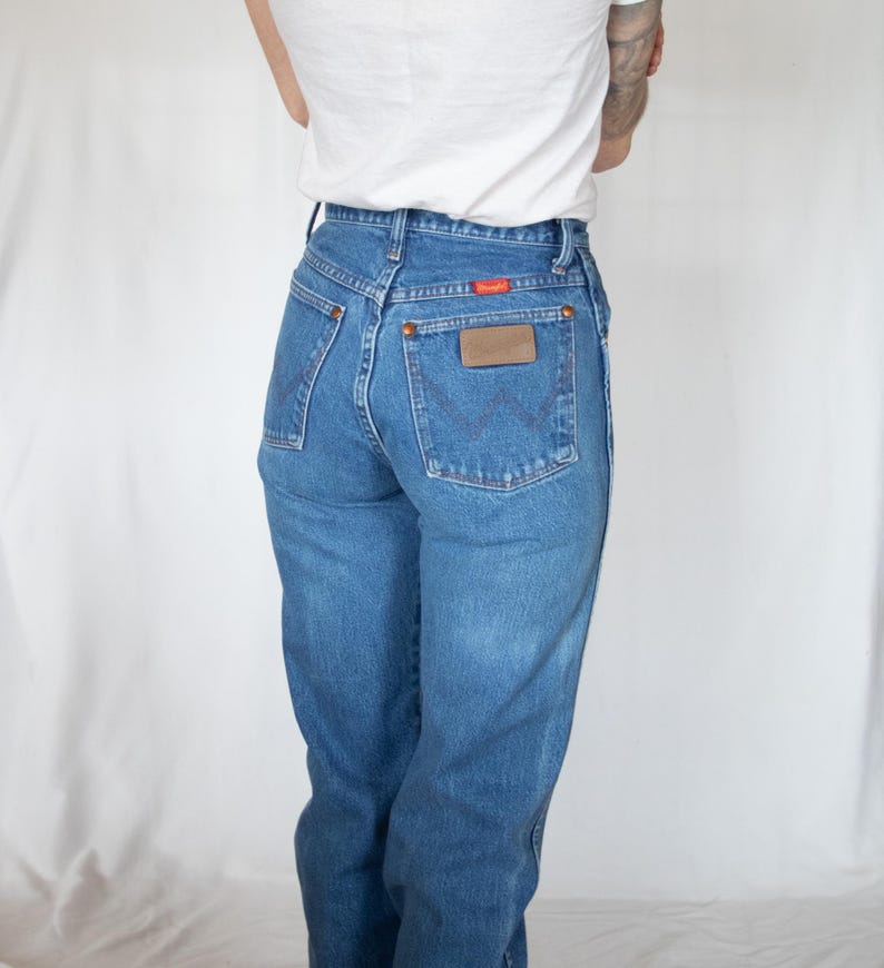 May include: High-waisted blue denim jeans featuring a classic five-pocket design, copper-toned rivets, and a brown leather patch on the back pocket. The back pocket displays a signature W-shaped stitching pattern and a small red Wrangler brand tag.