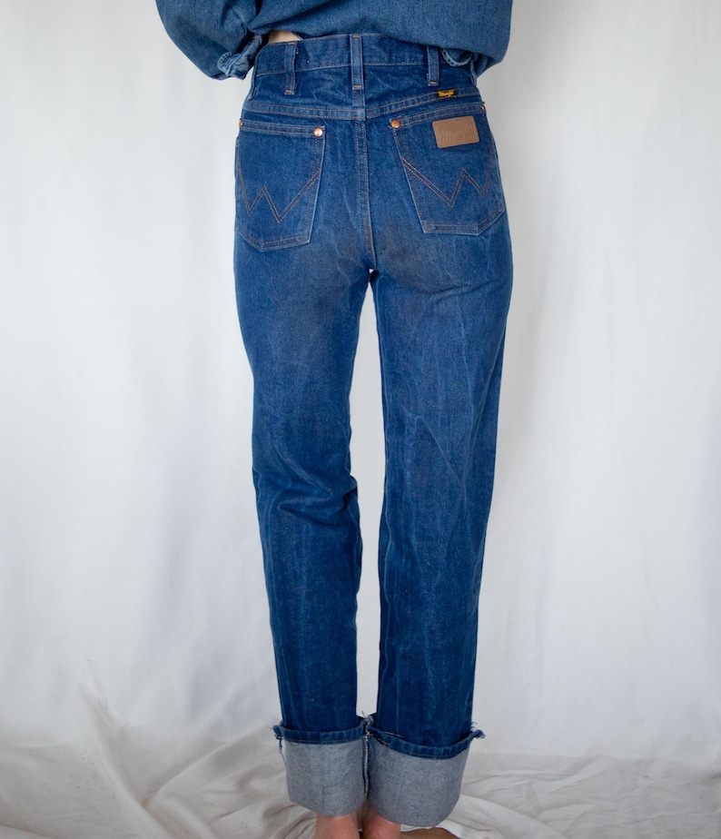 May include: A pair of dark blue denim jeans featuring a high-waisted fit, two back pockets with a signature W-shaped stitch pattern, copper-toned rivets, a brown leather patch on the right pocket, and wide cuffs rolled up at the bottom hem.