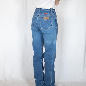 May include: Blue denim jeans featuring a high-waisted fit, a classic five-pocket design, and a brown leather patch on the back right pocket. The jeans include a small orange Wrangler brand tag and a signature W-stitch detail on the back pockets.