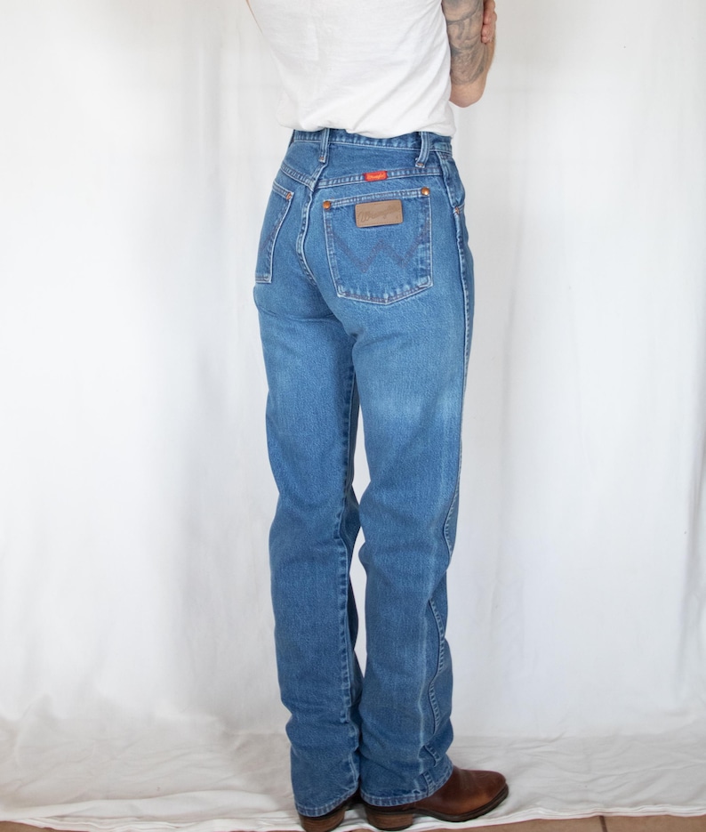 May include: Blue denim jeans featuring a high-waisted fit, a classic five-pocket design, and a brown leather patch on the back right pocket. The jeans include a small orange Wrangler brand tag and a signature W-stitch detail on the back pockets.