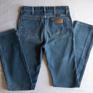 Puede incluir: Unos pantalones vaqueros azules con cintura alta y trabillas para cintur&oacute;n, dos bolsillos traseros con remaches de color cobre y un parche de cuero marr&oacute;n en el bolsillo derecho que muestra el nombre de la marca Wrangler en relieve.