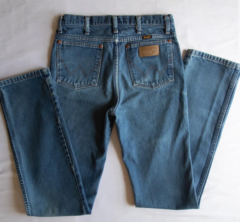 Puede incluir: Unos pantalones vaqueros azules con cintura alta y trabillas para cintur&oacute;n, dos bolsillos traseros con remaches de color cobre y un parche de cuero marr&oacute;n en el bolsillo derecho que muestra el nombre de la marca Wrangler en relieve.