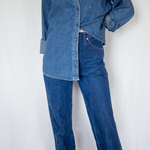 May include: A medium-wash blue denim button-down shirt with a chest pocket and long sleeves, paired with high-waisted dark blue denim jeans featuring a straight-leg cut and cuffed hems at the ankles, creating a coordinated denim-on-denim outfit.