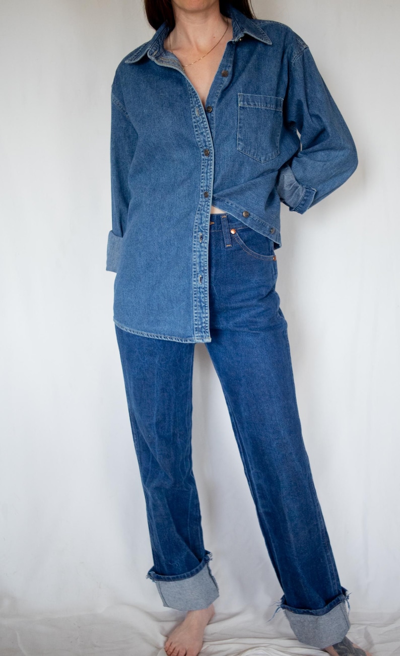 May include: A medium-wash blue denim button-down shirt with a chest pocket and long sleeves, paired with high-waisted dark blue denim jeans featuring a straight-leg cut and cuffed hems at the ankles, creating a coordinated denim-on-denim outfit.