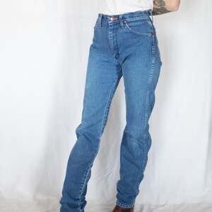 May include: High-waisted blue denim jeans featuring a classic five-pocket design, a button fly closure, and straight-leg cut. The jeans are paired with a plain white t-shirt and brown leather boots, showcasing a standard medium-wash denim finish.