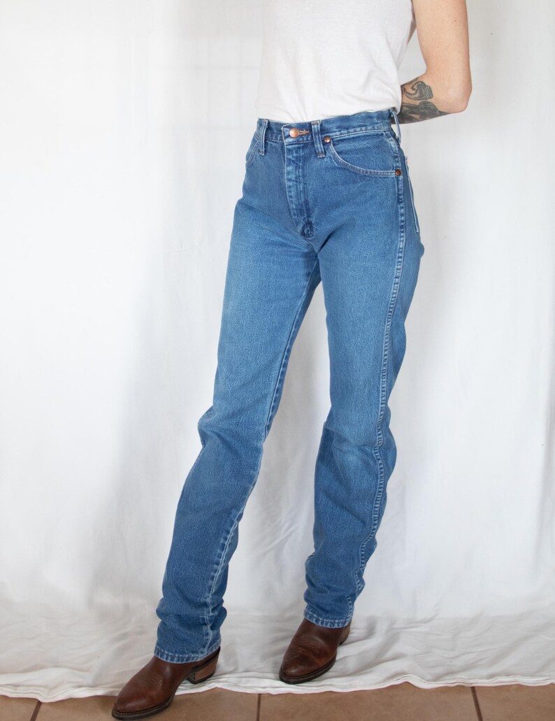 May include: High-waisted blue denim jeans featuring a classic five-pocket design, a button fly closure, and straight-leg cut. The jeans are paired with a plain white t-shirt and brown leather boots, showcasing a standard medium-wash denim finish.