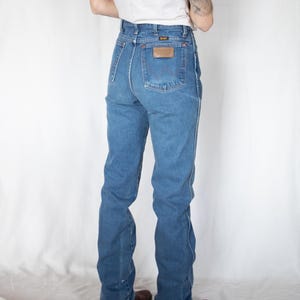 Puede incluir: Vaqueros de mezclilla azul de talle alto con corte recto, un bolsillo trasero con un parche de cuero marr&oacute;n de Wrangler y un dise&ntilde;o de costura en W. Los vaqueros incluyen trabillas para cintur&oacute;n, remaches cobrizos y cinco bolsillos.