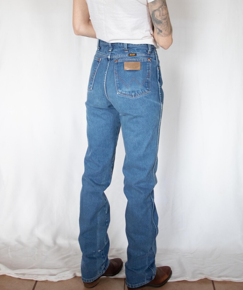 Puede incluir: Vaqueros de mezclilla azul de talle alto con corte recto, un bolsillo trasero con un parche de cuero marr&oacute;n de Wrangler y un dise&ntilde;o de costura en W. Los vaqueros incluyen trabillas para cintur&oacute;n, remaches cobrizos y cinco bolsillos.