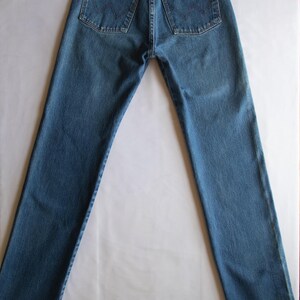 May include: A pair of blue denim jeans featuring a high-waisted design with a classic five-pocket construction. The back pockets display a signature W-shaped stitching pattern, and a brown leather-like patch with the Wrangler logo is attached.