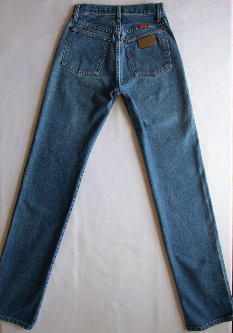 May include: A pair of blue denim jeans featuring a high-waisted design with a classic five-pocket construction. The back pockets display a signature W-shaped stitching pattern, and a brown leather-like patch with the Wrangler logo is attached.