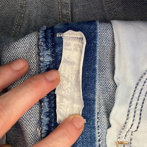 May include: A blue denim garment featuring a white fabric care label stitched onto the interior waistband. The denim fabric displays a classic twill weave texture with visible blue and white threads, and the label appears worn and faded.