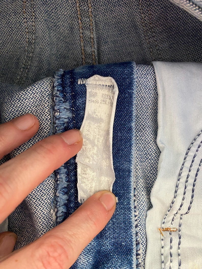 May include: A blue denim garment featuring a white fabric care label stitched onto the interior waistband. The denim fabric displays a classic twill weave texture with visible blue and white threads, and the label appears worn and faded.