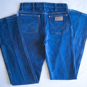 May include: A pair of blue denim jeans featuring a classic five-pocket design with copper-coloured rivets. The back pockets display a signature W-shaped stitching pattern in orange thread, and a brown leather-like patch with the Wrangler logo.